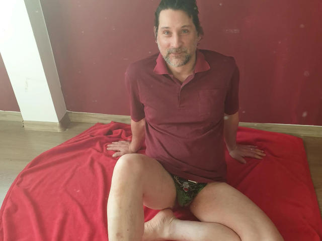 Mario41-hot, middle-aged, dark-haired, bearded, wearing maroon polo and green floral underwear, sitting on red blanket, legs spread, showing his hairy ass and flaccid dick.