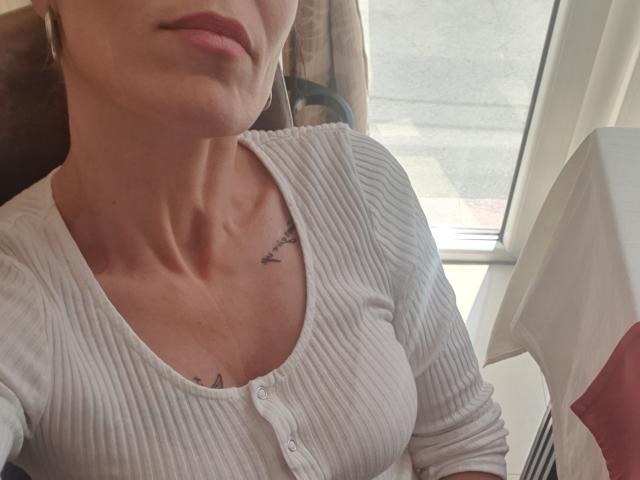 SweetMikyForYou's face is partially visible, wearing a ribbed white cardigan, showing small tits with a bird tattoo near the collarbone, sitting by a window.