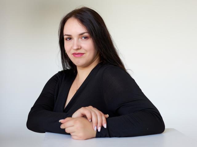 IsabelBlackberry has long black hair, wearing a black V-neck top, with pink lipstick, sitting with arms crossed, smiling subtly, and has small, cute tits.