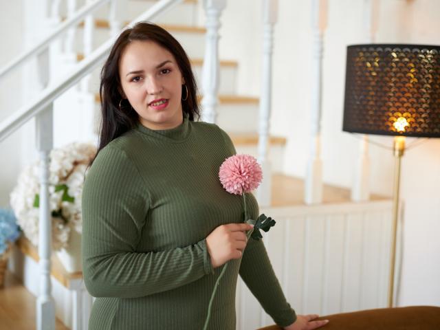 IsabelBlackberry with medium tits, green ribbed sweater, holding pink pom-pom flower, standing by white staircase, black lamp with warm glow, light wood floor, smiling, dark brown hair.