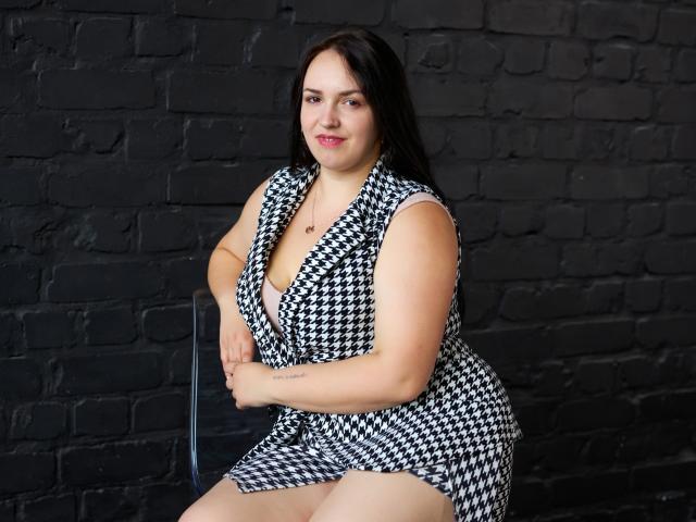IsabelBlackberry, de figura curvilínea con trasero grande y pechos pequeños, sentada en una silla, vestida con un vestido de cuadros de pied de poule, fondo de pared de ladrillo negro, cabello oscuro, lápiz labial rosa, brazos cruzados, ligera sonrisa..