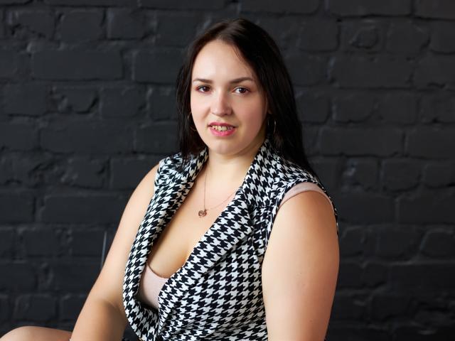 IsabelBlackberry with medium tits, wearing a black-and-white houndstooth dress, pale pink bra, dark hair, subtle smile, black brick wall, small necklace, sitting, slightly open dress.