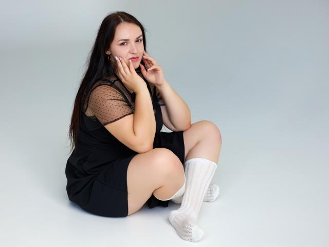 IsabelBlackberry with long brown hair, black dress with sheer polka-dot sleeves, white socks, sitting on the floor, hands near face, smiling, pink lips, soft light, cute.