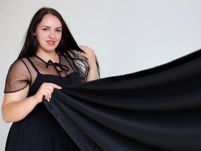 IsabelBlackberry with long black hair, wearing a sheer black polka-dot top, holding a black fabric, smiling, looking at the camera, showing her tits slightly through the fabric.