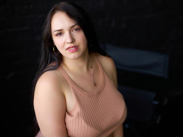 IsabelBlackberry, con cabello negro largo, un top rosa acanalado y aretes, sonriendo ligeramente, mostrando sus senos, lápiz labial rosa y piel clara, sobre un fondo oscuro..