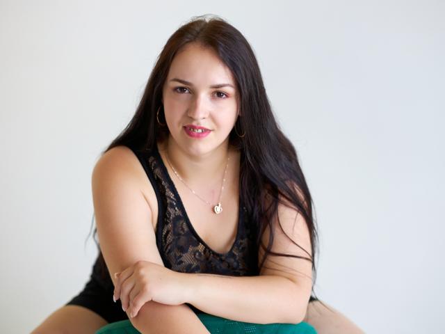 IsabelBlackberry has long black hair, wearing a black lace tank top, sitting with arms crossed, green chair, gold necklace, hoop earrings, smiling with pink lipstick, white background.