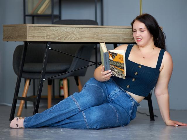 IsabelBlackberry, with dark hair, wearing a navy ribbed crop top with buttons and high-waisted blue jeans, sits on the floor reading a book, legs crossed.