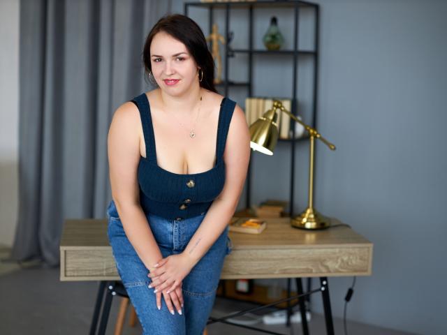 IsabelBlackberry, curvy with big tits, wears a dark blue tank top and blue jeans, sitting on a wooden desk, hands on knees, smiling, brown hair, gold lamp behind.