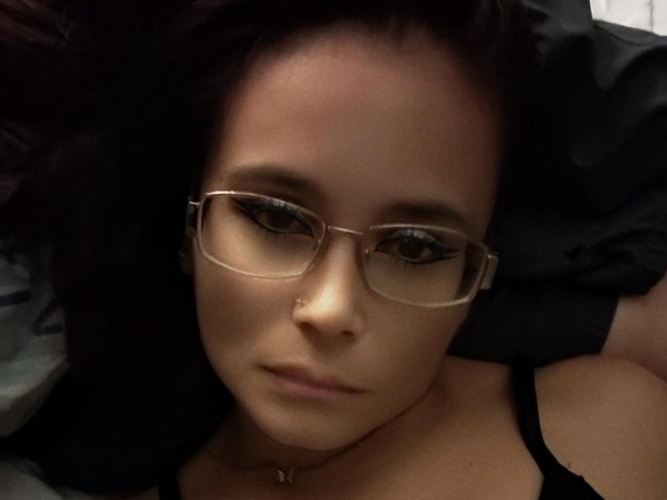 BabeDolly69s has dark hair, glasses, and a nose ring, lying down with a serious expression, wearing a black top, showing a subtle cleavage.