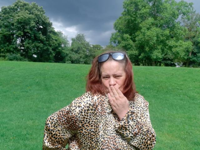 Kathy75-hot with red hair, wearing a leopard print shirt, has her hand over her mouth, sunglasses on her head, standing on green grass, dark cloudy sky in background.