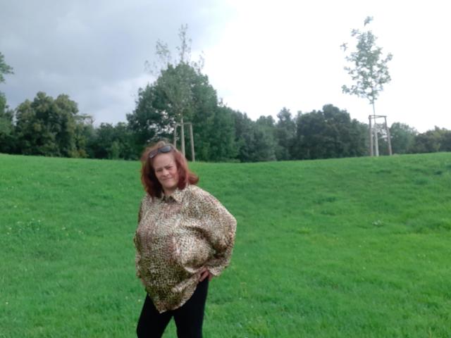 Kathy75-hot stands in a grassy field, wearing a patterned shirt, black pants, and a smile, with her hands on her hips, and her red hair blowing slightly.