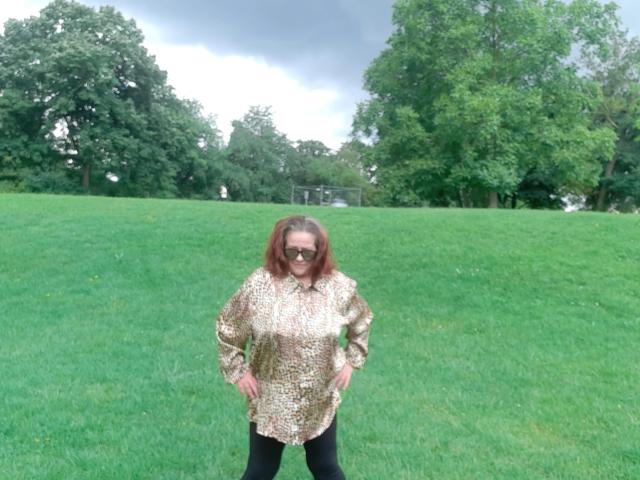Kathy75-hot with red hair, wearing a gold patterned shirt, black pants, and sunglasses, stands with hands on hips in a grassy field, trees in the background.