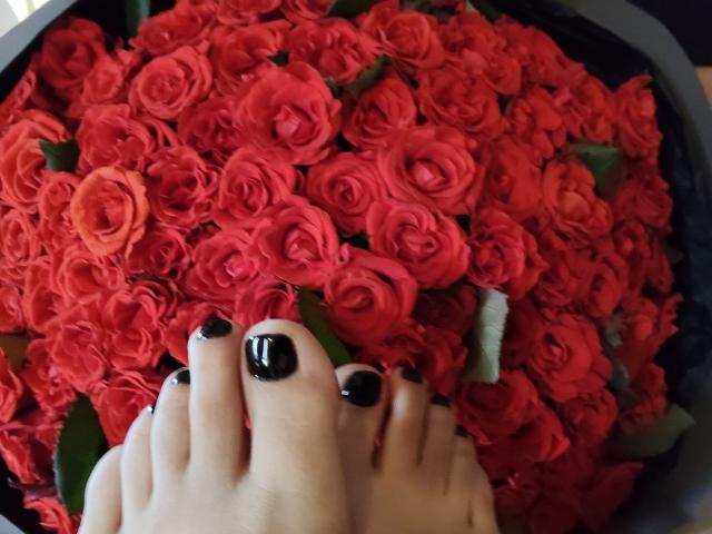 Nurri's voet, met glanzende zwarte nagellak, rust op een grote bos rode rozen. Haar tenen zijn lichtjes gekruld, waardoor haar gepolijste teentjes zichtbaar zijn..