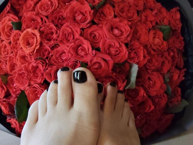 I piedi di Nurri, con le unghie dipinte di nero, riposano su una grande ciotola piena di rose rosse. Le dita dei piedi sono leggermente incurvate, mostrando lo smalto nero lucido..