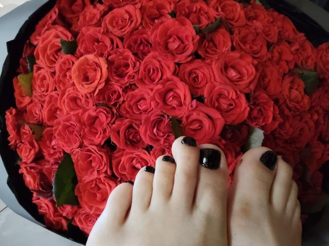 I piedi di Nurri, con le unghie dipinte di nero, si trovano nell'angolo in basso a destra, accanto a un grande bouquet circolare di rose rosse che riempie un vaso nero..