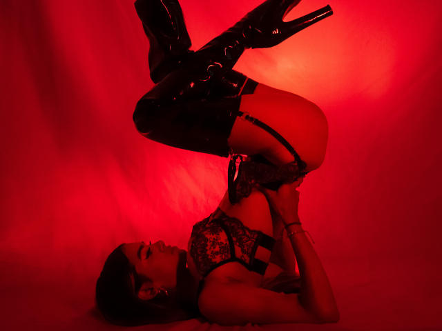 VanelopeRusso in black lace lingerie and thigh-high boots, doing a headstand with legs up, showcasing her butt and high heels. Red lighting.