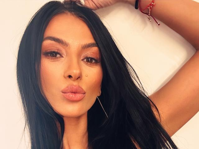 KinkyAmelie with long black hair, brown eyes, and full pouty lips, wearing a red bracelet, has a small beauty mark below her nose, and is looking at the camera.