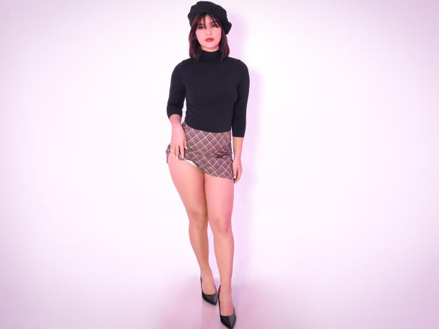 ScarlletZhen with black turtleneck, plaid mini-skirt, black hat, and black heels. She has a curvy butt, long legs, and is standing with a slightly parted lip.
