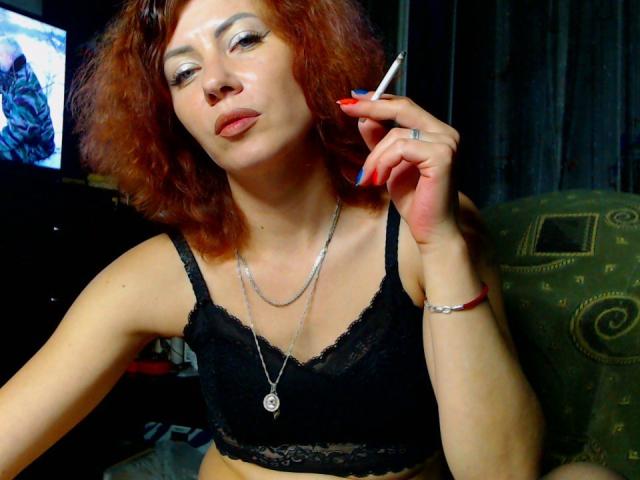 JuliaIce with red curly hair, wearing a black lace bra, smoking a cigarette, has shiny silver eyeshadow, red nails, and a silver necklace, sitting on a green chair.