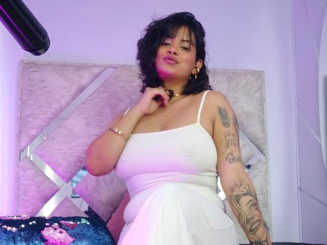 MariGonz, avec ses cheveux noirs et ondulés, portant une robe blanche, a de grandes seins et un bras tatoué, et se tient dans une pièce éclairée en violet, tout en se touchant le menton..