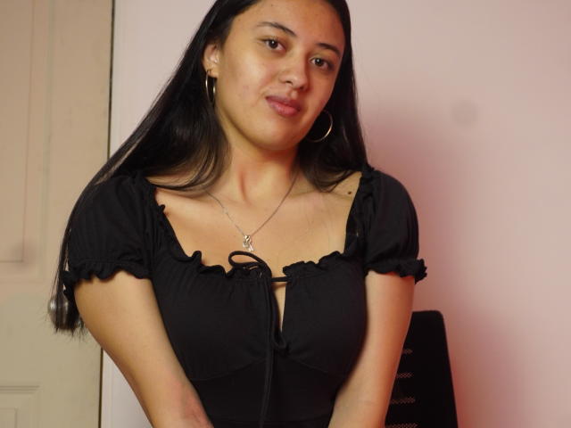 MiaBloom69 with long black hair, wearing a black ruffled top, small tits, hoop earrings, and a silver necklace, smiling softly at the camera.