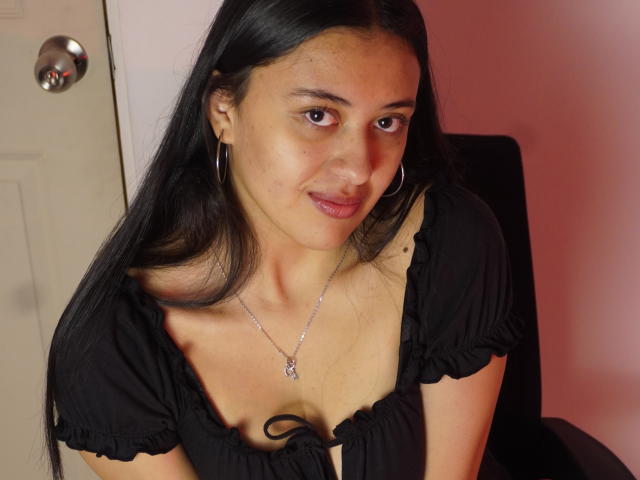 MiaBloom69 with long black hair, hoop earrings, and a black top showing her small tits and a necklace, smiling slightly, sitting in a chair.