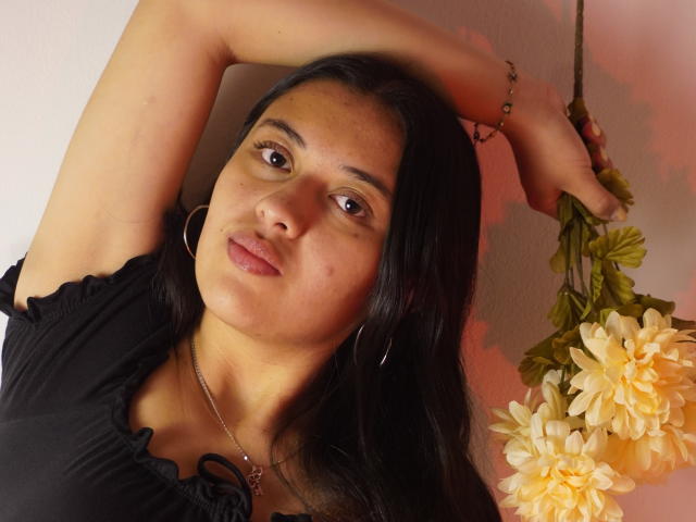 MiaBloom69 with black hair, brown eyes, and medium tits, wearing a black top, hoop earrings, and necklace, holding flowers, arms raised, and a relaxed, slightly smirking expression.
