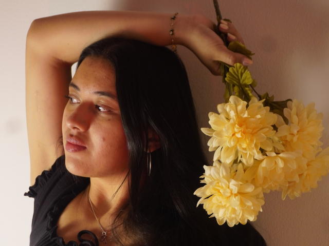MiaBloom69 with dark hair, holding yellow flowers, arm raised, wearing black top, hoop earrings, and necklace, looking side, soft light, slightly pouty, subtle makeup, tan skin.