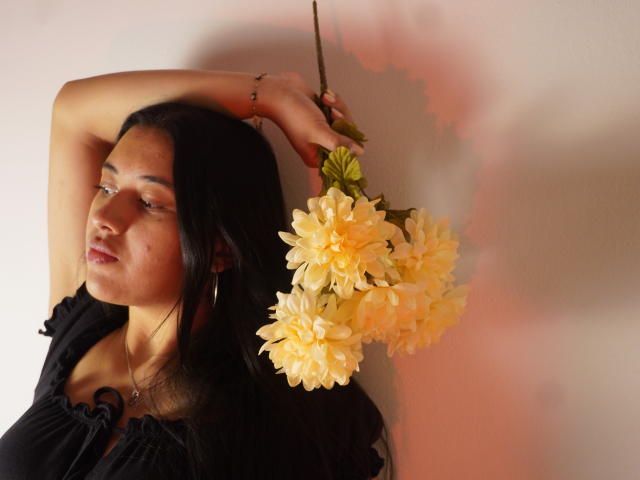 MiaBloom69 with dark hair, black top, and hoop earrings, holding yellow flowers above her head, arms raised, looking sideways, soft lighting, pink shadow on right.