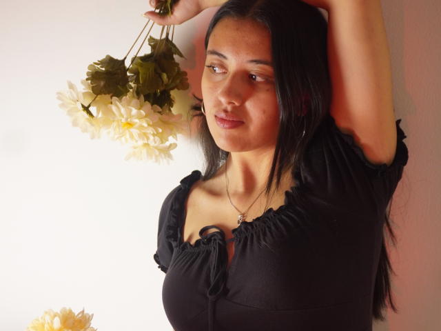 MiaBloom69 with black dress, arms raised, holding white flowers, soft light, medium tits, long black hair, brown eyes, slight smile, white wall, subtle jewelry, relaxed pose.