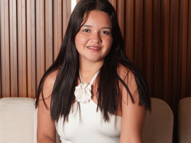 SaraJhonsson with long black hair, smiling, wearing a white halter top with a flower, has brown eyes, and a medium build, sitting on a beige couch.