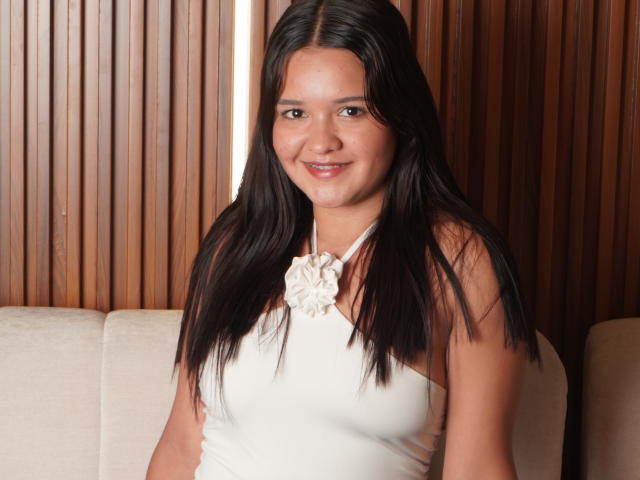 SaraJhonsson has long dark hair, a cute smile, and wears a white sleeveless top with a flower accessory, showing small tits. She sits on a beige couch.