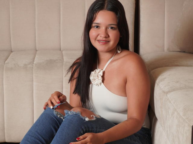 SaraJhonsson with dark hair, medium tits, wearing a white halter top, blue ripped jeans, sitting on a beige couch, smiling at the camera, red nails, and a white necklace.