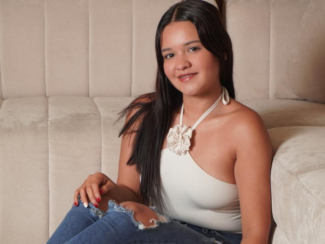 SaraJhonsson with long black hair, wearing a white halter top with a flower, and ripped blue jeans, sitting on a beige couch, smiling softly at the camera.