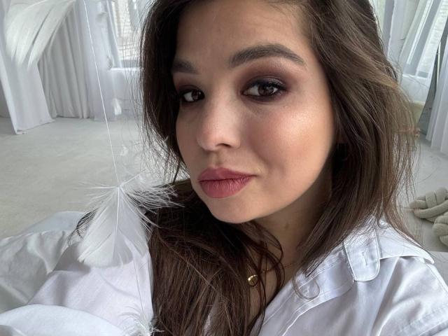 ALisaKissy with brown eyes, pink lipstick, and white shirt holding a feather, looking at camera with a slight smile. She has long brown hair and a necklace.