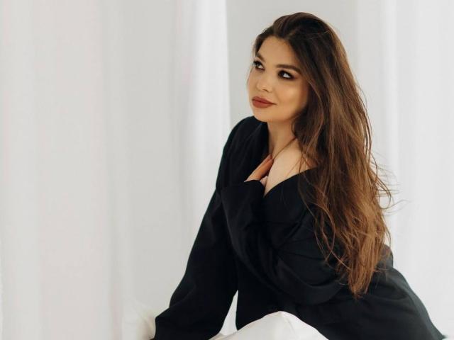 ALisaKissy with long brown hair, wearing a black top, sitting on white fabric, looking over her shoulder with a subtle smile, her tits partially covered by her hand.