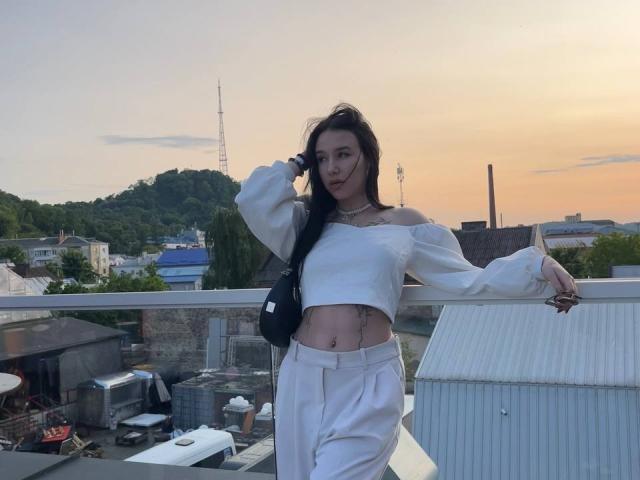 NikkiGoldX stands on a rooftop, wearing a white off-shoulder crop top and high-waisted pants, with a black bag, exposing her flat stomach and a small belly button piercing.