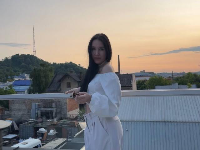 NikkiGoldX with long black hair, off-shoulder white top, and white pants, standing on a rooftop at sunset, holding a phone, with a calm, slightly serious expression.