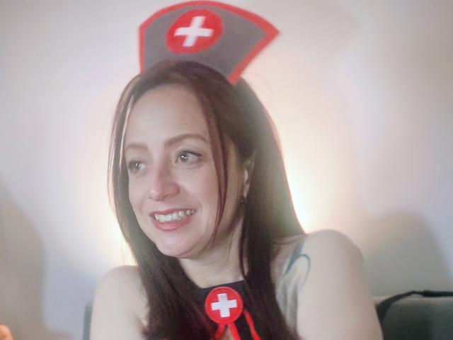 CatalinaMonyque with long brown hair, smiling, wearing a nurse hat with a red cross, black top, and red button, showing her bare shoulders and small tattoos.