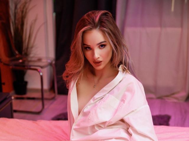 HeySiry with long blonde hair, wearing a white, slightly open, silky robe, exposing her tits, looking seductively at the camera, in a pink-lit bedroom.