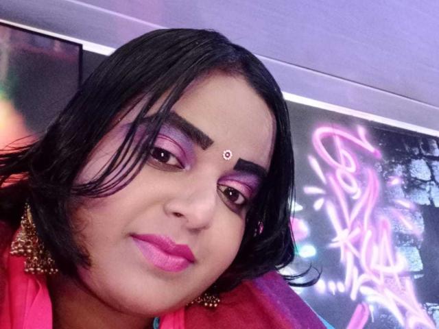 SusanaEshwarCurvy has dark hair, purple eyeshadow, pink lipstick, and a bindi on her forehead. She's wearing a pink top with gold earrings, looking at the camera.