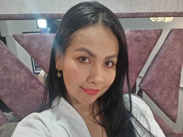 MartinaCrox with long black hair, light skin, red lipstick, and small gold earrings, smiling at the camera, wearing a white top, showing her pretty face and cute ass.