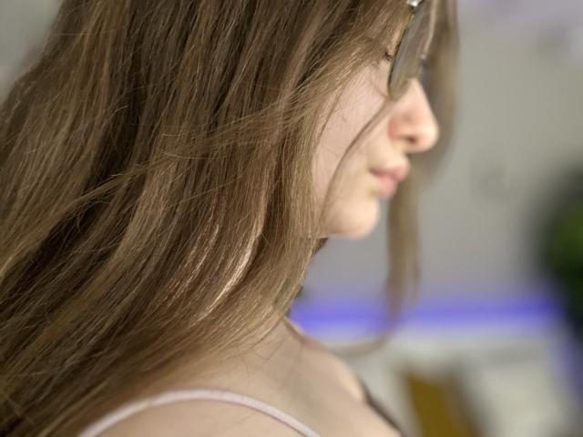 MagikPeachh's side profile with long brown hair, wearing glasses, showing a delicate nose and pinkish lips, in a soft pink top, blurred background.