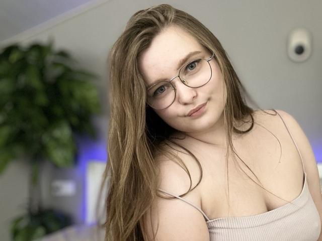 MagikPeachh with long brown hair, glasses, and a slight smile, wearing a beige ribbed top that shows her tits, sitting in a softly lit room.