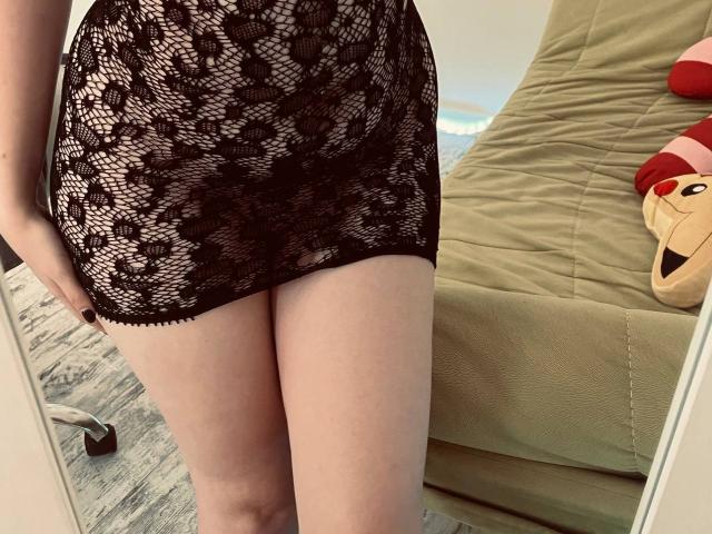La gonna in pizzo di Valissia è nera e trasparente, rivelando le sue gambe chiare e un po' del suo sedere. È in piedi accanto a una sedia verde con un peluche..