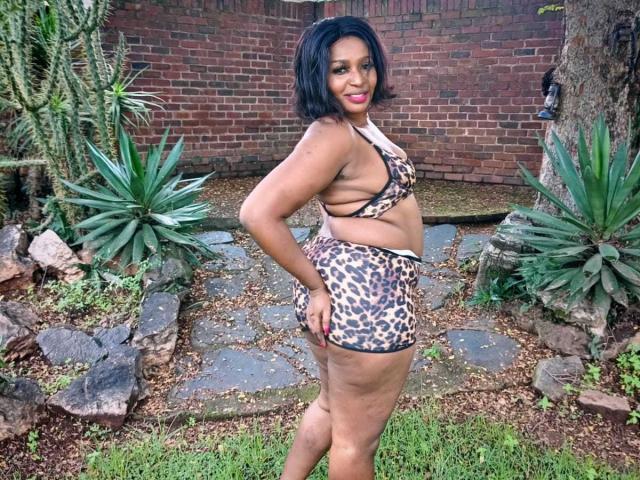 HotXBums with short black hair, brown skin, and curvy ass wearing leopard print bikini, standing sideways, hands on hips, looking back with a smile. Outdoor garden setting.