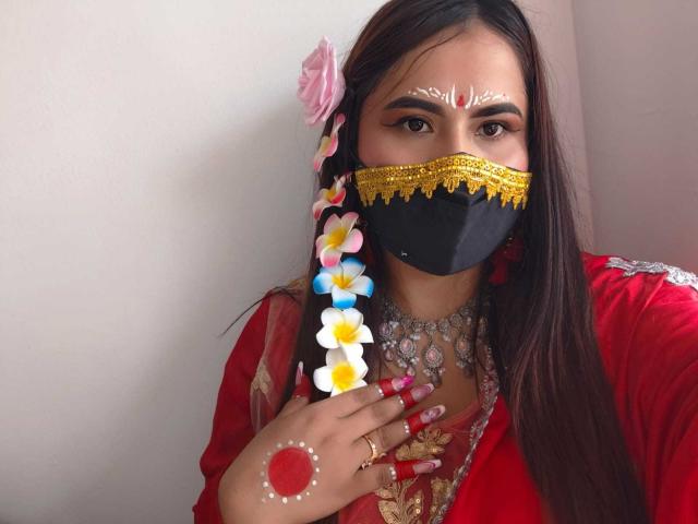 Dalia Dubey, con lunghi capelli castani, indossa un abito rosso, una maschera dorata e nera, una ghirlanda di fiori, fiori bianchi e gialli, un braccialetto rosso e una collana d'argento, e guarda verso la telecamera..
