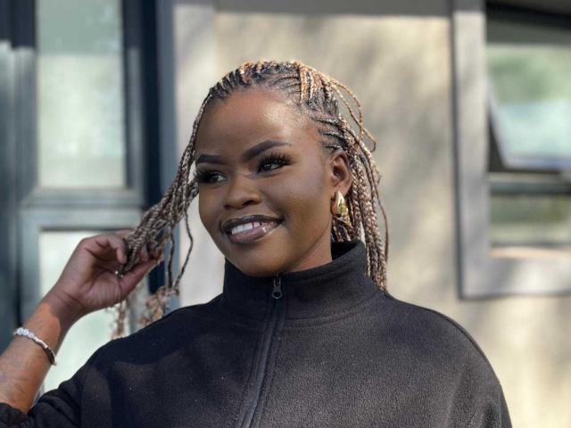 JessicaGreenX with dark skin, braided hair, gold earrings, black zip-up jacket, smiling, holding hair, shiny makeup, silver bracelet, standing outside, sunny day.