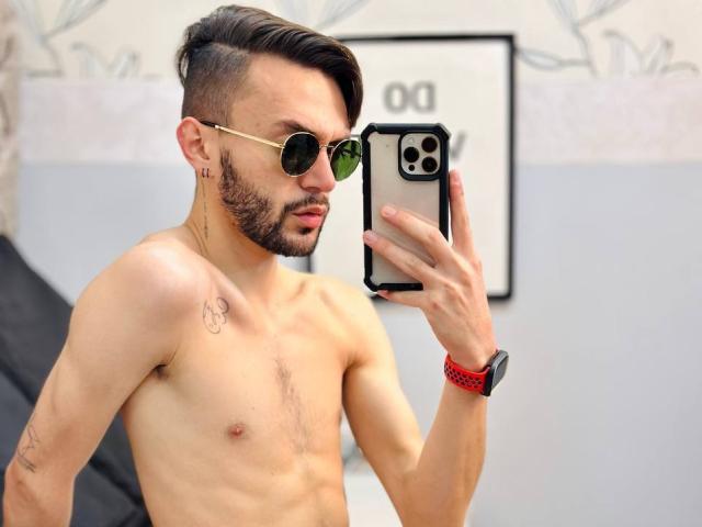 JakeMagnus ohne Hemd, mit einer trainierten, behaarten Brust, trägt grüne Aviator-Sonnenbrillen und macht ein Selfie mit einem iPhone. Rotes Armband, Kurzhaarschnitt mit Iro und ein Tattoo an seiner rechten Schulter..