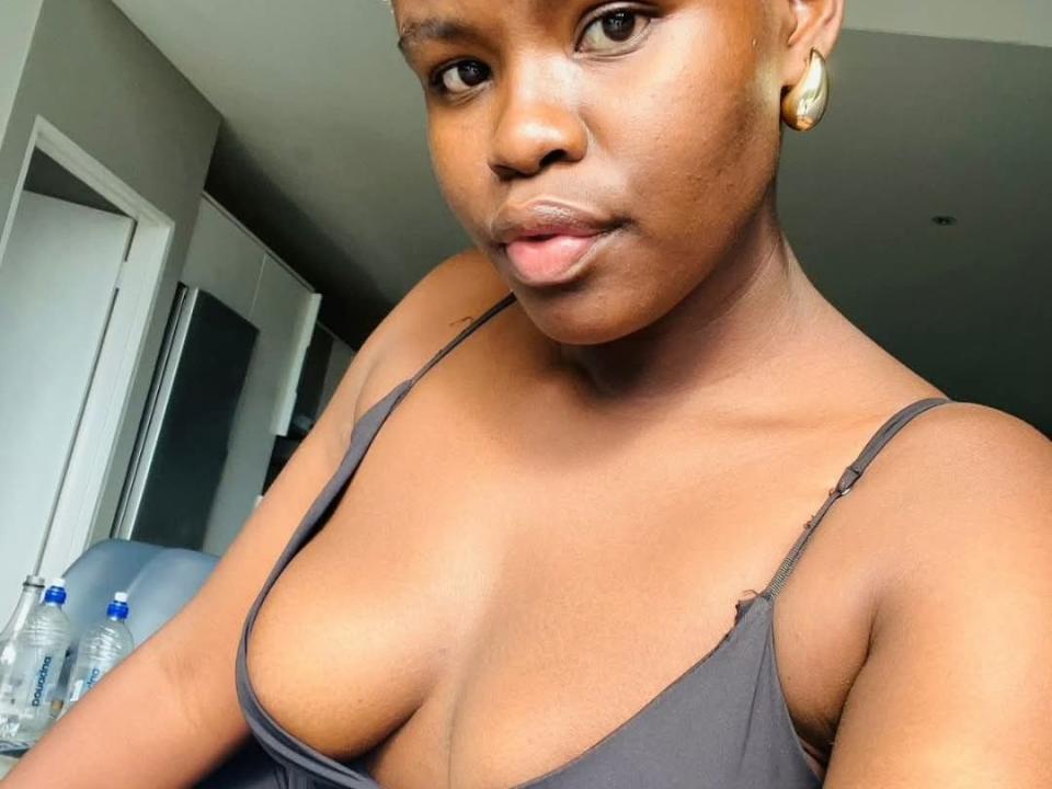 JessicaGreenX has dark brown skin, gold earrings, and wears a black spaghetti-strap top showing her tits. She's looking at the camera with a slight pout. Water bottles are in the background.